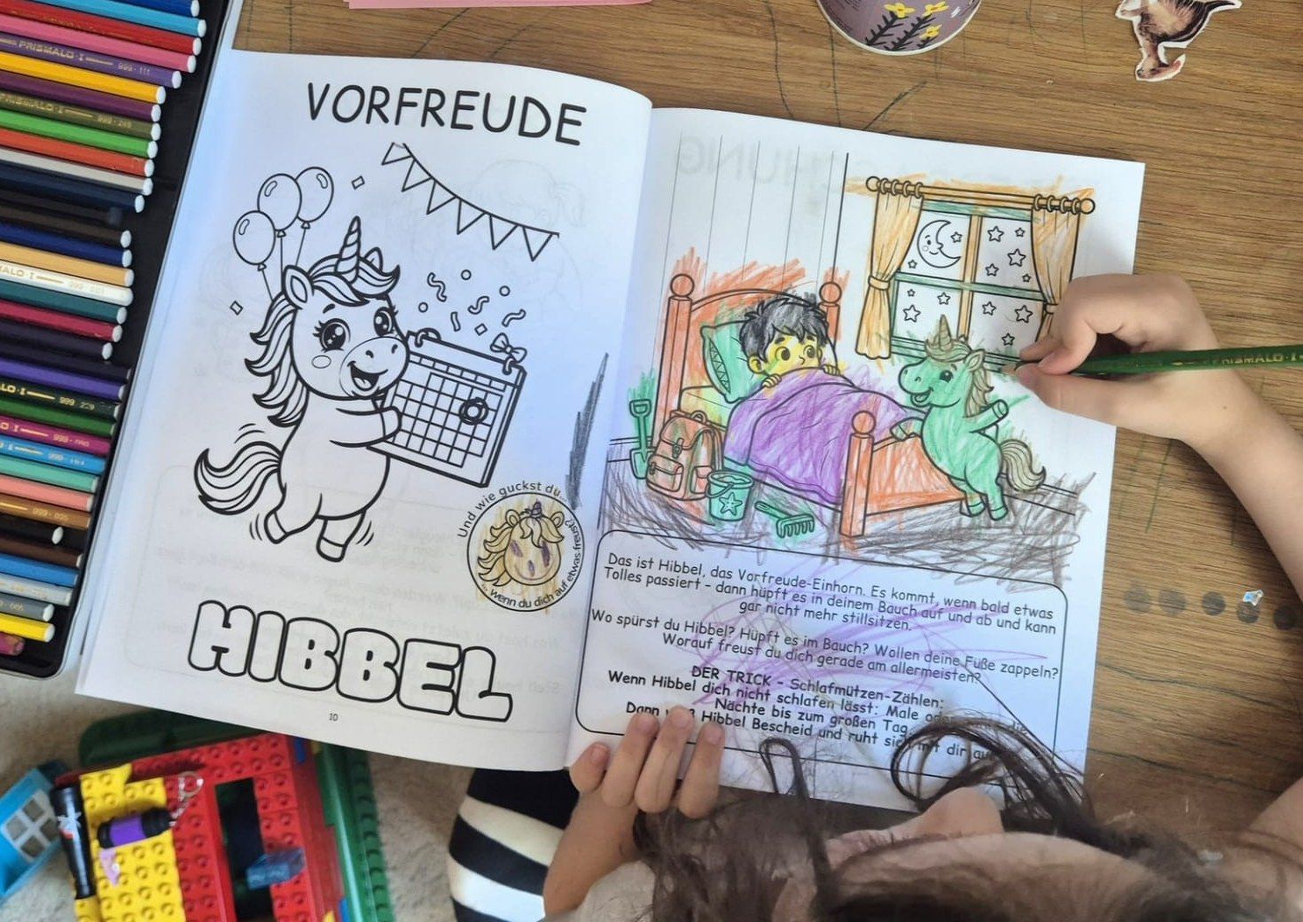 Ein Kind malt in den Gefühlseinhörnern die Doppelseite Vorfreude mit Buntstiften aus; links das Einhorn Hibbel, rechts eine Szene mit einem Kind im Bett.